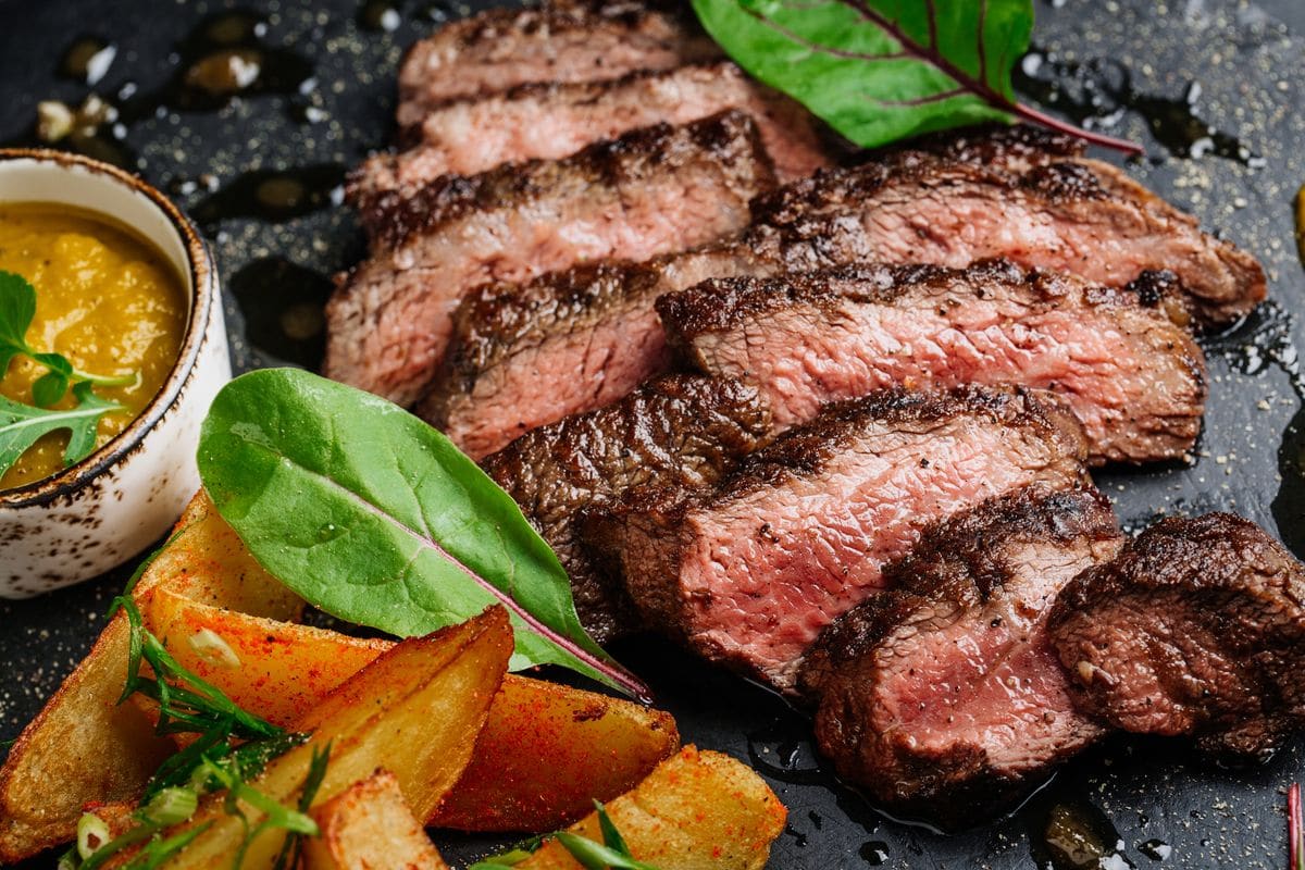 Sliced grilled steak with potatoes and leaves on a dark plate